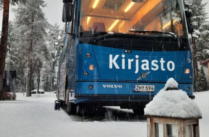 Kirjastoauto Kotikujan Konsta lumisessa maisemassa. Etualalla puinen lyhty.